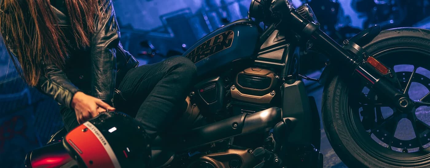 A woman is shown sitting on a blue 2022 Harley-Davidson Sportster S parked in a garage.