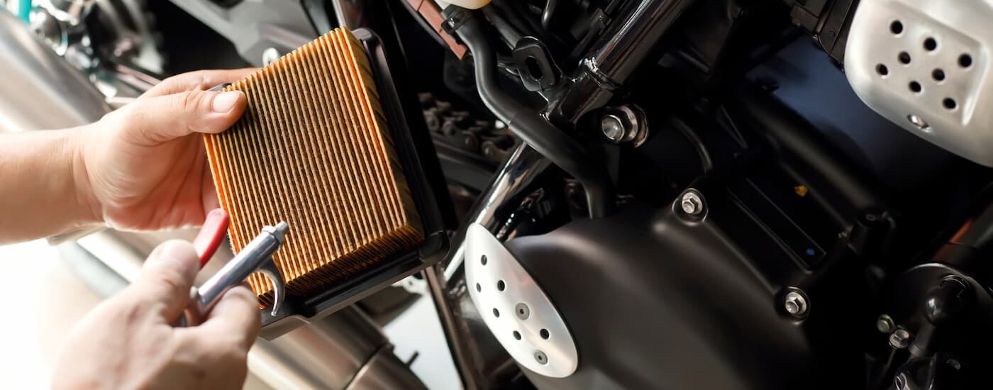 A mechanic is shown using an air compressor to clean a dirty motorcycle air filter.