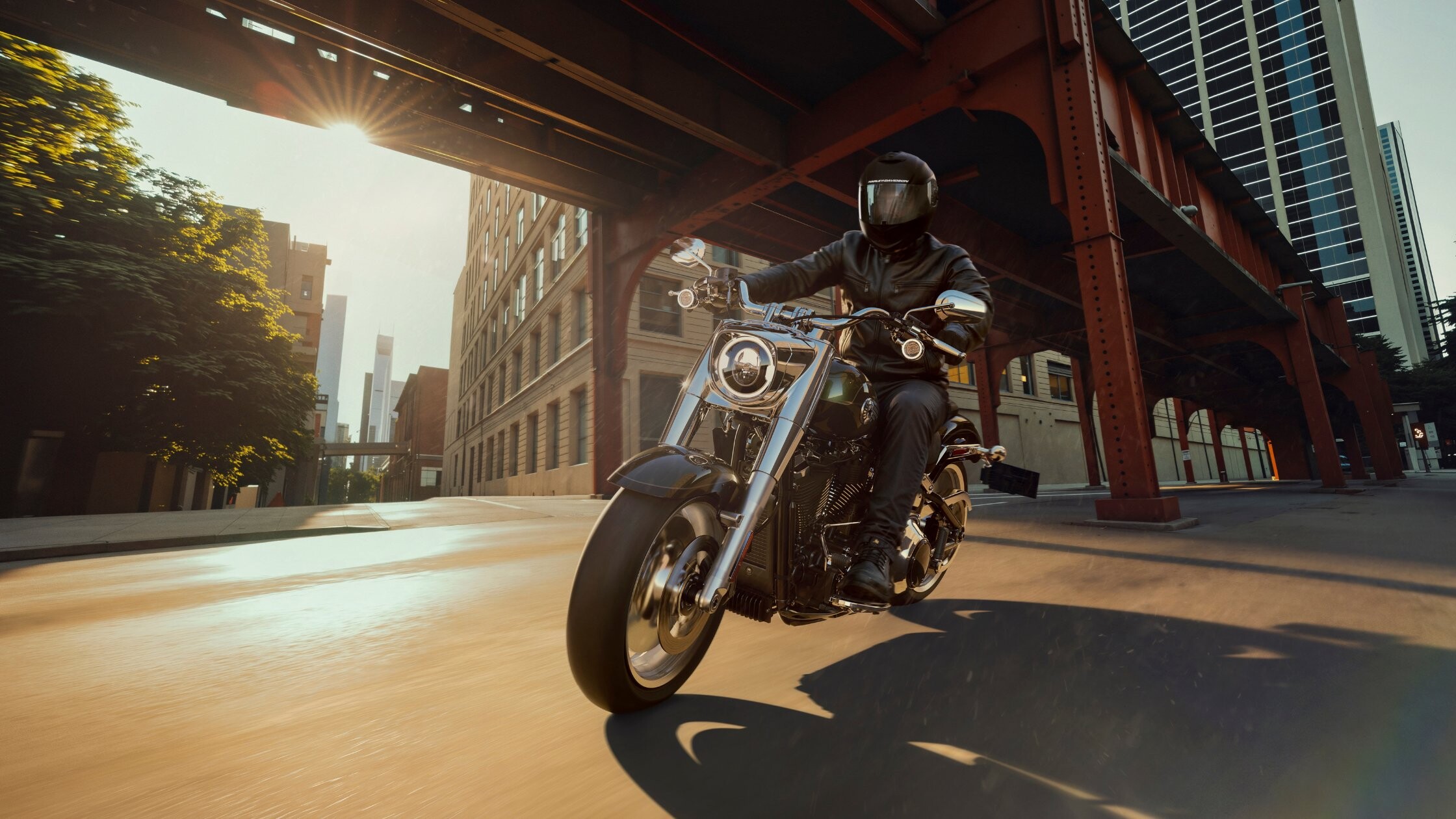 Rider cruising 2026 Harley-Davidson® Fat Boy® through city streets beneath elevated rail bridge