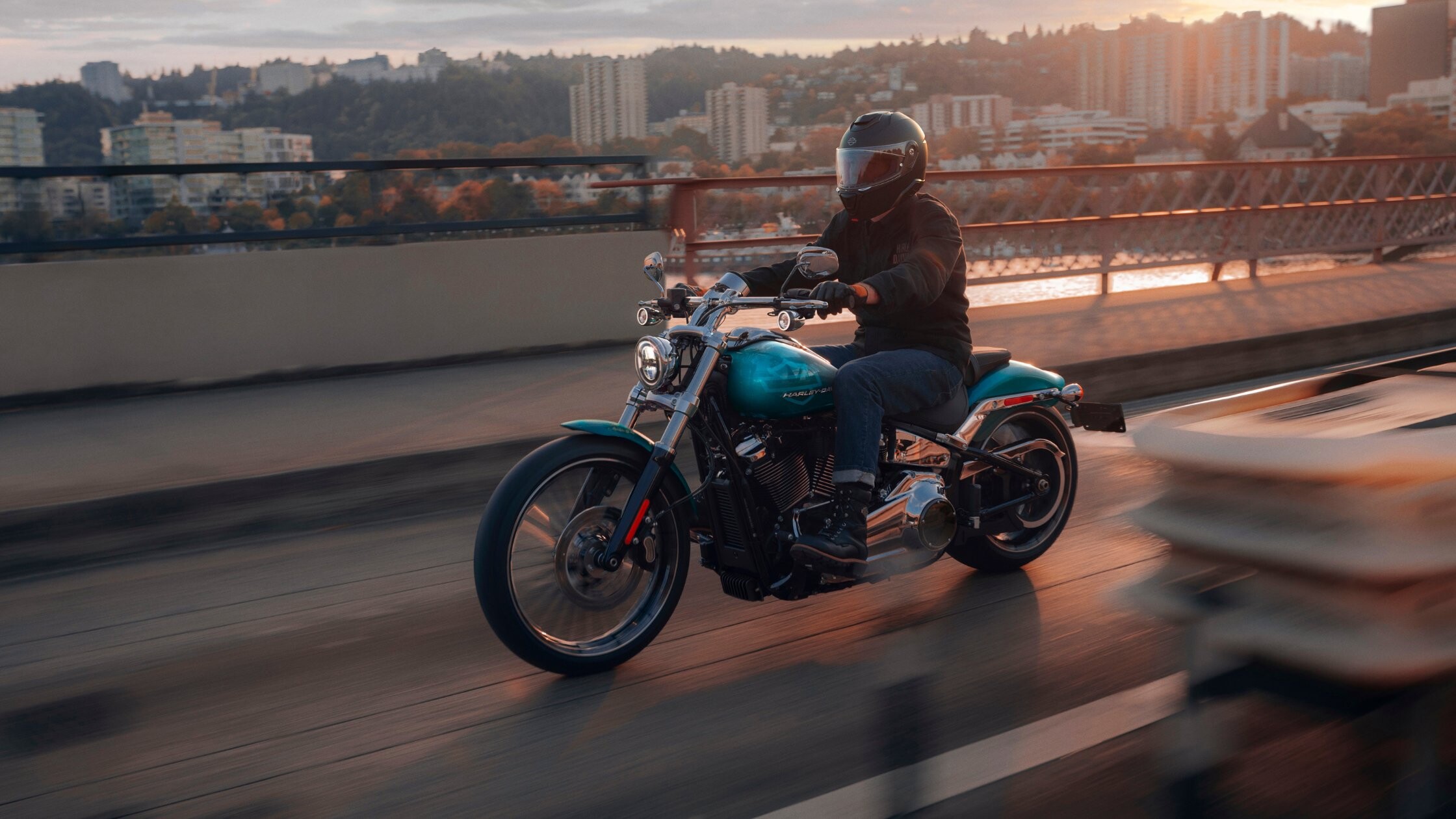 Teal 2026 Harley-Davidson® Breakout® cruising city bridge at sunset with urban skyline
