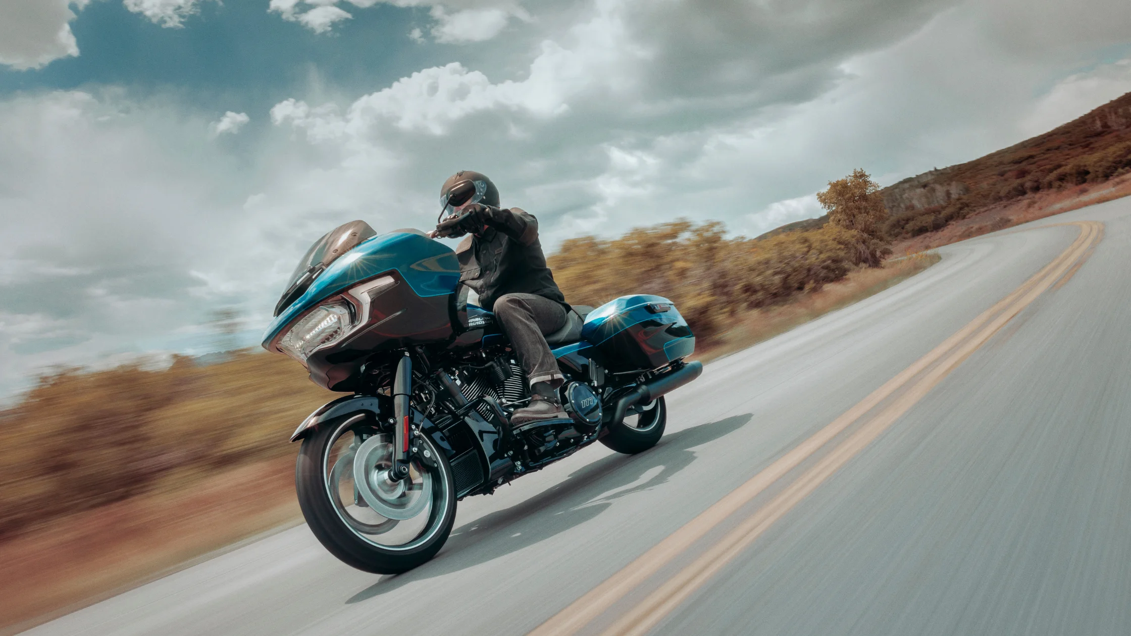 Rider leans a blue 2026 Harley-Davidson® Road Glide® through a curving open road under clouds