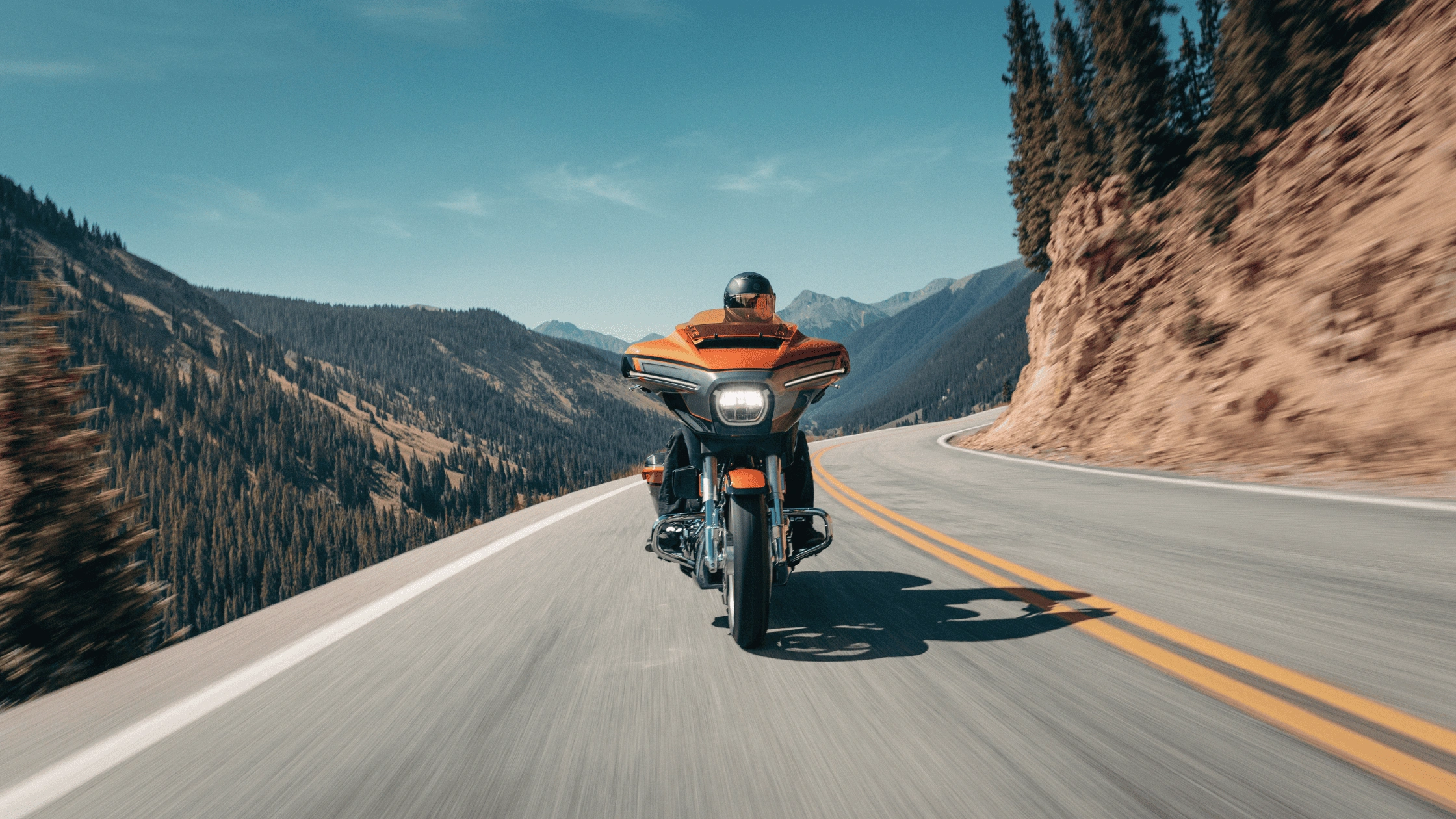 Rear view of a rider on a 2026 Harley-Davidson CVO Street Glide in orange navigating a winding mountain highway, available in Baton Rouge LA